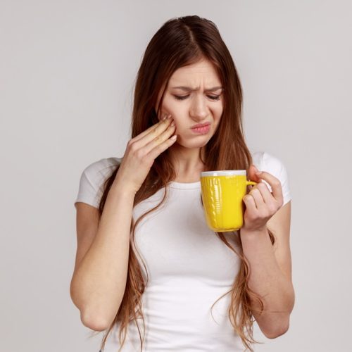 Sick unhealthy woman touching chin suffering tooth ache, has sensitive teeth, feels pain after drinking cold water, wearing white T-shirt. Indoor studio shot isolated on gray background.