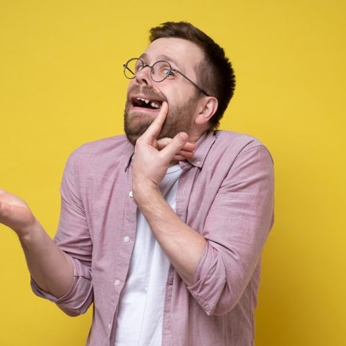 Perplexed man in glasses and casual clothes opened his mouth, shows a missing tooth and makes an inquiring gesture with hand. Isolated, yellow background.