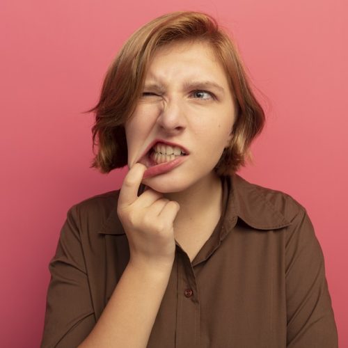 frowning young blonde girl looking at side pulling her bottom lip with one eye closed isolated on pink background