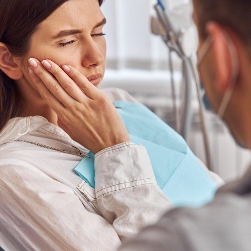 Young woman having toothache, sitting in a dental chair at the clinic. Female patient with terrible toothache visiting dentist